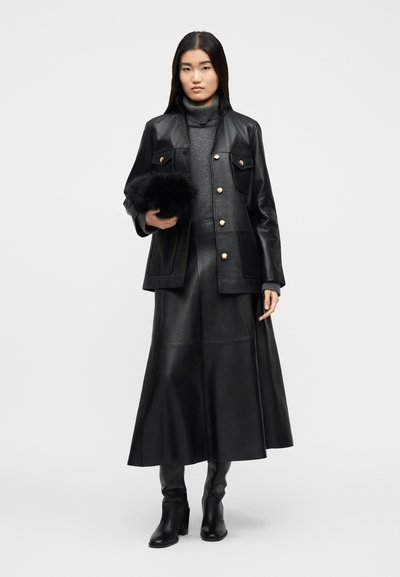 Γυναίκα που φοράει μαύρο δερμάτινο μπουφάν και φούστα, γκρι πουλόβερ με ψηλό λαιμό, μαύρες μπότες με τακούνι, κρατώντας μικρό τσαντάκι με μαύρη γούνα σε λευκό φόντο.