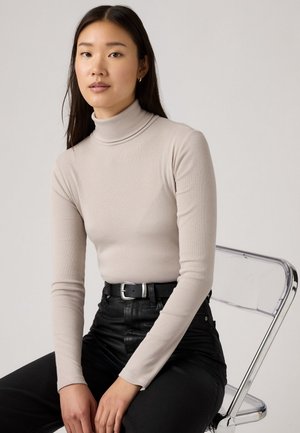 Beige gestreifter Rollkragenpullover mit langenÄrmeln, kombiniert mit hoch taillierten schwarzen Hosen und einem schwarzen Gürtel mit einer silbernen Schnalle, sitzt auf einem transparenten Stuhl.