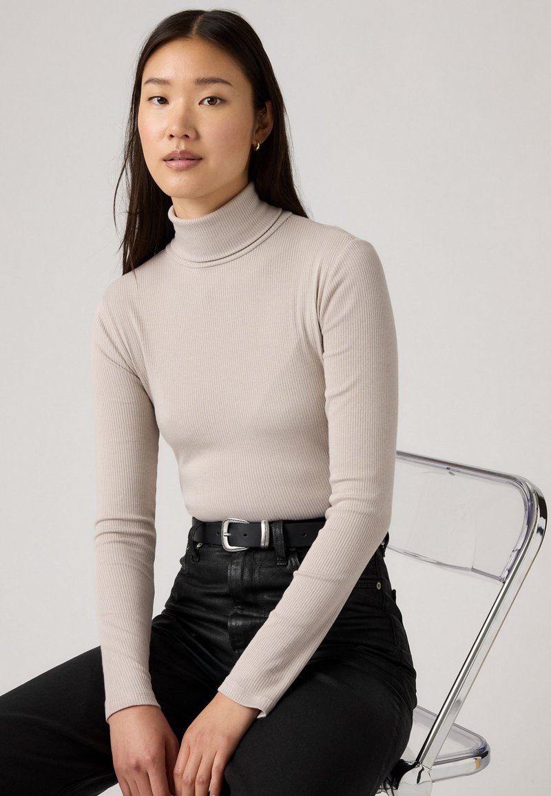 Beige gestreifter Rollkragenpullover mit langenÄrmeln, kombiniert mit hoch taillierten schwarzen Hosen und einem schwarzen Gürtel mit einer silbernen Schnalle, sitzt auf einem transparenten Stuhl.