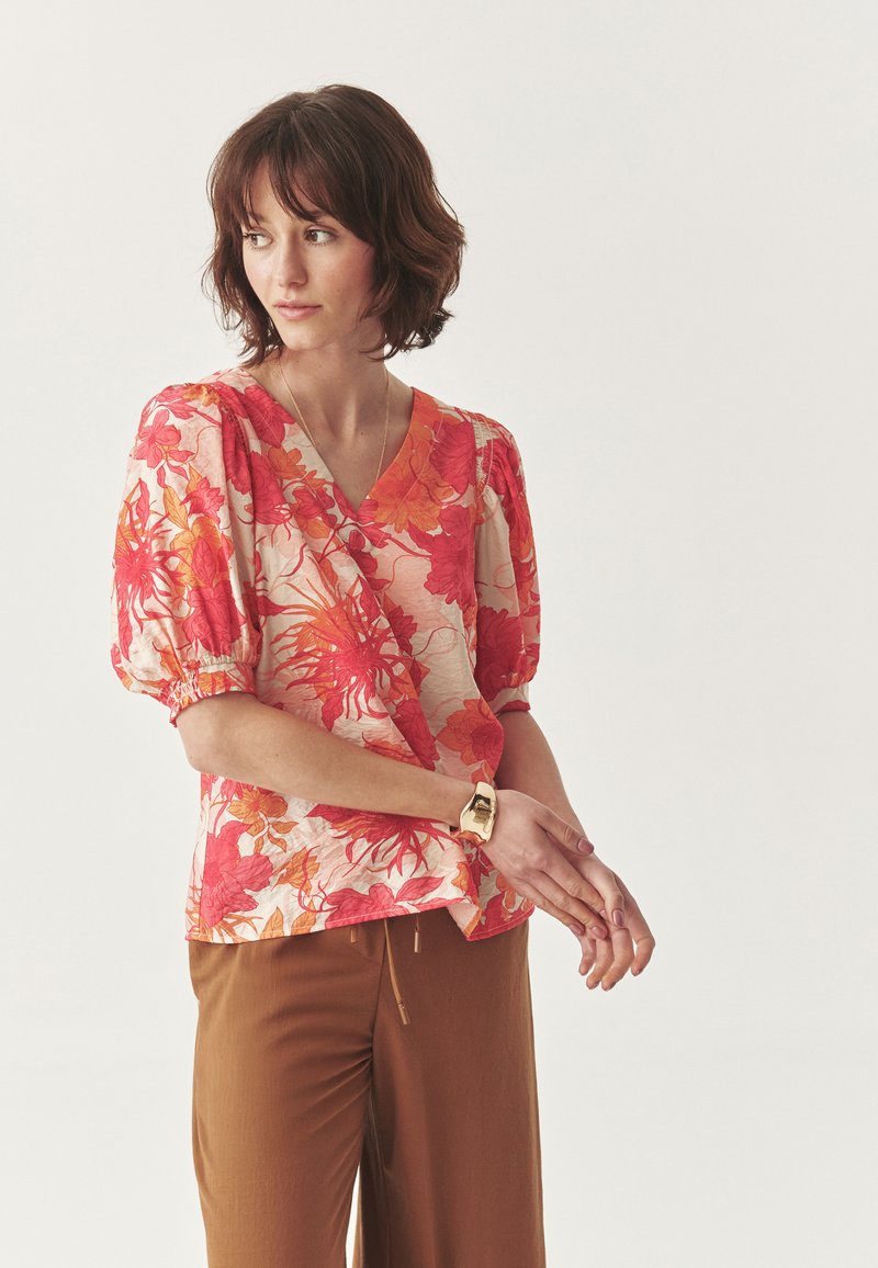 Blumenwickelbluse in Rosatönen und Orange, mit Puffärmeln, V-Ausschnitt und strukturiertem Stoff, kombiniert mit braunen Hosen.