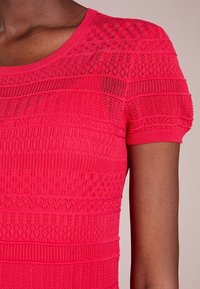 Červený pletený top s texturovanými vzory a přiléhavým střihem, krátkými rukávy a kulatým výstřihem. Materiál vypadá lehký a prodyšný.