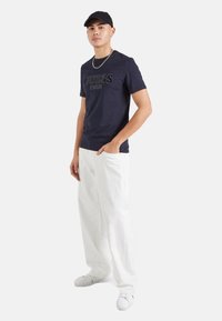 Camiseta de algodón azul marino con el logo "GUESS", combinada con pantalones blancos holgados y zapatillas blancas, llevada con una gorra negra y una cadena de plata.