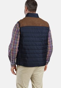 Marineblaue Steppweste mit braunen Wildlederschultern und gestepptem Design. Trägt man über einem mehrfarbigen Karobluse, die einen lässigen Schichtlook zeigt.