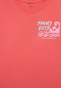 Korallivärvi puuvillane T-särk, millel on sädelevas hõbedases tekst "SUNNY DAYS" ja laine graafika, sileda tekstuuri ning klassikalise ümara kaelusega disain.