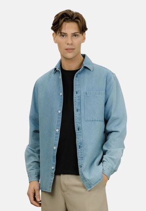 Ung mann iført en lys blå denimskjorte over en svart t-skjorte og beige bukser, stående med den ene hånden i lommen mot hvit bakgrunn.