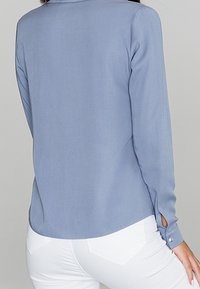 Femme portant un chemisier bleu clair à manches longues avec poignets boutonnés et un pantalon blanc, vue de dos sur un fond uni.