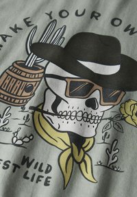 Camiseta gráfica con un cráneo que lleva gafas de sol, un sombrero de vaquero y una bandana amarilla, con cactus decorativos y rosas sobre un fondo verde claro.