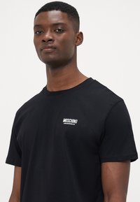Juoda medvilninė marškinėliai su klasikiniu apvaliu kaklu ir balta išsiuvinėta logo kairėje krūtinėje, lygi tekstūra ir trumpomis rankovėmis.
