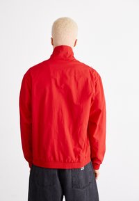 Röd lättviktsjacka med hög krage, elastisk midja och långa ärmar. Har en slät, matt textur och inga synliga logotyper.