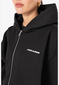 Čierna mikina s kapucňou na zips vyrobená z bavlnenej zmesi, s priestrannou kapucňou, predným zipsom a bielym logom "PEGADOR" na ľavej hrudi.
