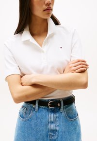 Woman wearing white polo shirt and blue jeans with black belt, arms crossed, facing slightly right with neutral expression.
