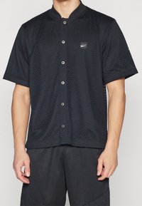 Maglietta nera in rete a maniche corte con chiusura a bottoni, dotata di colletto rotondo e piccolo logo Nike. Tessuto texturizzato con una vestibilità ampia.