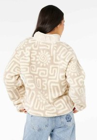 Beige fleecejakke med hvide geometriske mønstre, høj krave og en blød tekstur. Bemærkelsesværdigt soldesign på ryggen og afslappet pasform.
