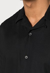 Camisa negra de manga corta con cuello clásico, textura sutil y dos botones en el frente. La tela parece suave y ligera.