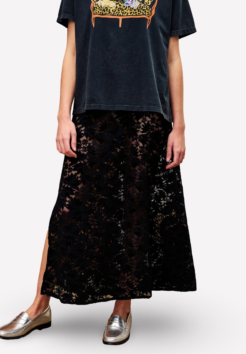 Schwarzer Midirock aus floraler Spitze mit seitlichem Schlitz, kombiniert mit einem verblassten schwarzen T-Shirt mit Grafik und metallic-silbernen Loafers, getragen von einer Person.