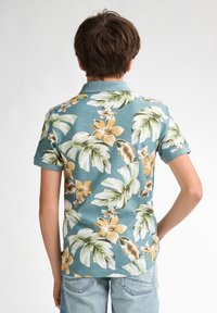Koszulka polo z krótkim rękawem w tkaninie w odcieniu teal, zdobiona tropikalnym wzorem kwiatowym z dużymi zielonymi liśćmi i pomarańczowymi kwiatami. Model z kołnierzykiem.