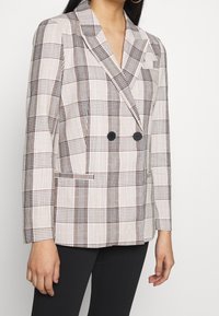 Blazer doppiopetto in un tessuto beige chiaro con motivo a quadri marroni, bottoni neri e colletto a scialle; include tasche laterali.