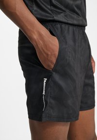Svarta sportshorts med randig design, försedd med sidfickor och en vit logotyp på nedre vänstra benet. Lättvikts tygkänsla.