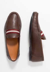 Chaussures à enfiler en cuir marron avec une finition texturée, un accent rayé rouge et blanc sur le cou-de-pied, et une semelle en caoutchouc noire plate.