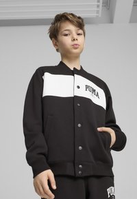 Czarna i biała kurtka PUMA z zapięciem na guziki, ściągaczami na rękawach i bocznymi kieszeniami. Posiada szeroki biały pasek i logo. Miękka tkanina.
