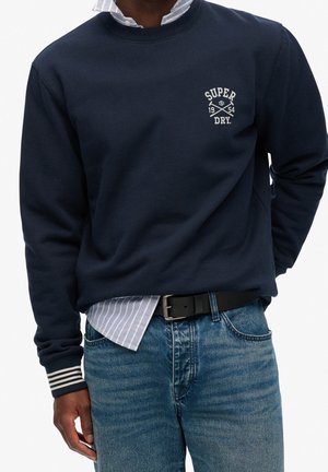 Mężczyzna w granatowej bluzie z logiem "Super Dry 1954", w paski mankietach, pod spodem w paskowaną koszulę, niebieskie jeansy i czarny pasek.