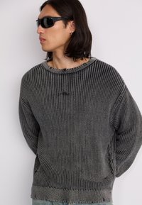 Knit sweater in gray with ribbed texture, wide crew neck, and loose fit. Worn with black sunglasses, showcasing modern design.
