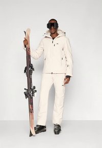 Άντρας φορώντας μπεζ χιονοδρομική στολή και μαύρα γυαλιά χιονοδρομίας, κρατώντας ένα ζευγάρι σκι κάθετα, στέκεται μπροστά σε απλό φόντο.