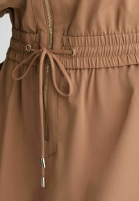Tessuto marrone con una superficie liscia, dotato di una cerniera dorata, cintura elastica e chiusura con cordino e punte metalliche.