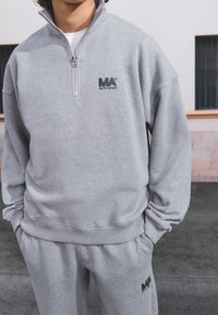 Sudadera gris y pantalones a juego en tela suave. Presenta un cuello con cremallera y un logo en negro en el pecho y la pierna del pantalón.