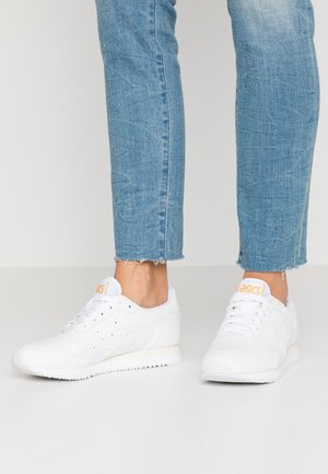 Zapatillas blancas Asics usadas con vaqueros cortos azul claro sobre un fondo blanco liso.