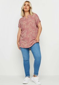 Blusa corta a maniche corte rosa con motivo floreale, colletto a punta e orlo asimmetrico, abbinata a jeans skinny blu e sneakers bianche.