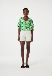 Groene bloemenblouse met pofmouwen, gecombineerd met crèmekleurige shorts en zwarte sandalen. Opvallende patronen zijn witte bloemen en blauwe accenten.