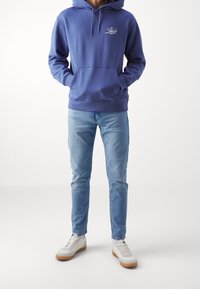 Man i blå Levi's hoodie, ljusblå jeans, vita sneakers, står mot en plain vit bakgrund med händerna i hoodien.