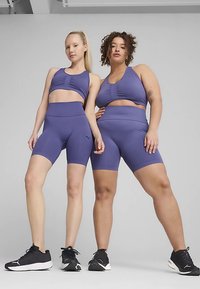 Μοβ σετ αθλητικής συλλογής που περιλαμβάνει εφαρμοστά αθλητικά σουτιέν και σορτς. Το ύφασμα φαίνεται λείο με διακριτική ριμπ υφή και λογότυπα ως διακοσμητικά στοιχεία.