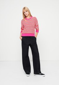 Punase ja valge triibulise kampsuniga, millel on roosa ääristus, kombineeritud tumedate laiade pükstega. Modell seisab neutraalsel taustal, käed taskutes.