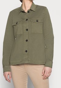 Olivově zelená košile na knoflíky s dvěma náprsními kapsami, černými knoflíky a límečkem. Vyrobena z hladké, texturované látky.