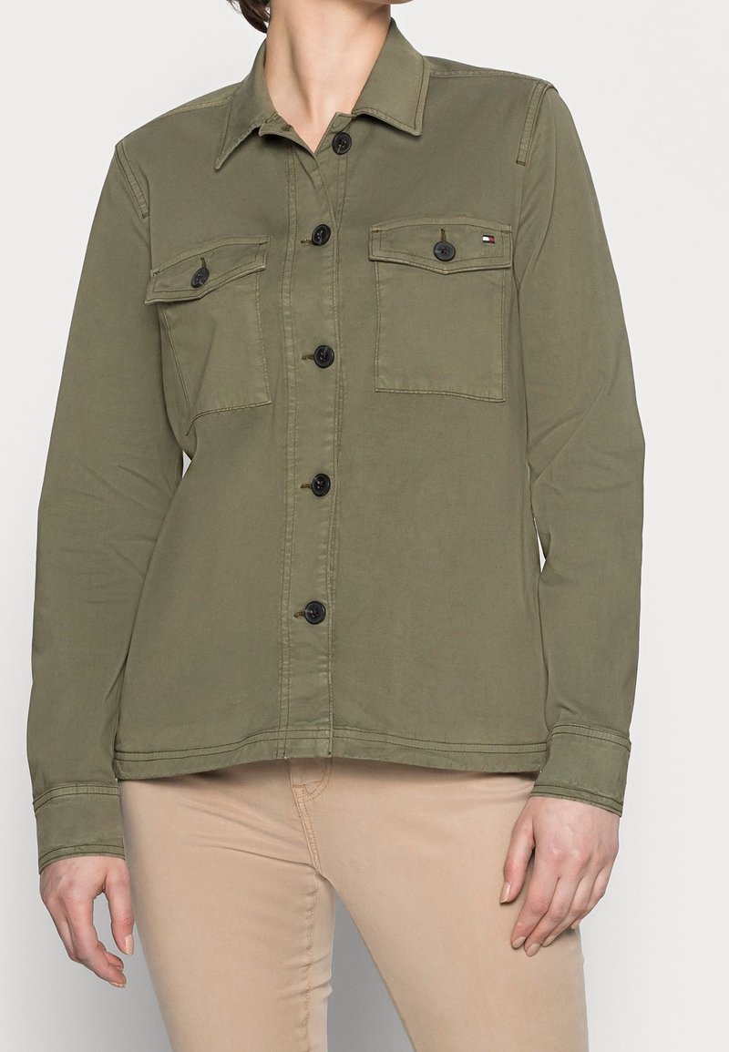 Olivově zelená košile na knoflíky s dvěma náprsními kapsami, černými knoflíky a límečkem. Vyrobena z hladké, texturované látky.