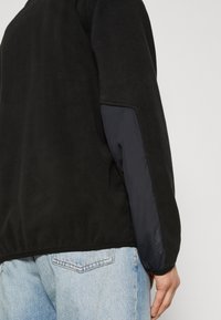 Pullover in pile nero con corpo strutturato, maniche lisce blu navy ed orlo elastico, abbinato a jeans di denim blu chiaro.