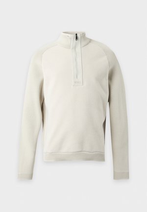 Un homme porte un pull crème à col zippé sur une chemise blanche, un jean foncé et des mocassins noirs, sur un fond clair et uni.