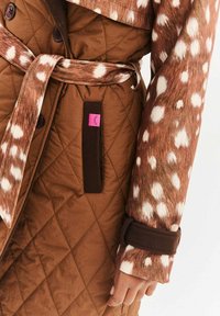 Brązowy pikowany płaszcz z rękawem w cętkowany wzór, pasującym paskiem do wiązania oraz różową naszywką z logo po boku. Miękka faktura, zapięcie na guziki.