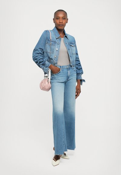 Woman wearing a cropped denim jacket, light gray top, high-waisted wide-leg faded jeans, white pointed heels, and carrying a small pink purse.