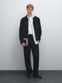 Junger Mann trägt eine schwarze Jacke, ein weiß karierter Hemd, schwarze Hose, hält Notizbücher und steht vor einer schlichten grauen Wand.