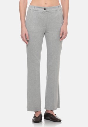 Persona che indossa pantaloni grigio chiaro a gamba dritta con bottone frontale e scarpe marroni scure senza lacci, in piedi davanti a uno sfondo semplice.