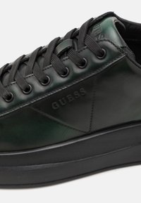 Sneakers in pelle nera con dettagli verdi, punta tonda, lacci neri e suola in gomma elevata. Presenta il logo "GUESS" in rilievo sul lato.