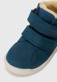Chaussure en cuir teal avec deux bandes Velcro, semelle en caoutchouc et doublure en polaire. Présente un détail en caoutchouc sur le bout et un logo embossé.