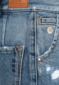 Tela de mezclilla azul clara que presenta un bolsillo trasero, costuras naranjas en contraste y un acento de estrella cerca de un botón. Logotipo de Scotch & Soda visible.