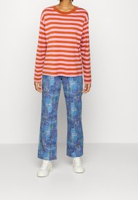 Osoba v červeném a růžovém pruhovaném tričku s dlouhým rukávem, modrých kalhotách s motivem očí a bílých vysokých teniskách na jednobarevném pozadí.