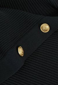 Materialul negru cu textura ribbed prezintă două butoane rotunde aurii, manoperite cu un emblema detaliată, evidențiind textura și cusăturile decorative.