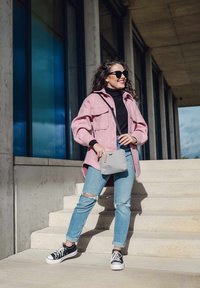 Světle růžová oversized bunda přes černý rolák, modré roztrhané džíny a černé tenisky. Drží malou šedou tašku. Slunné venkovní prostředí.