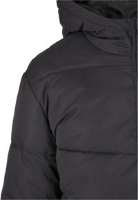 Veste d'hiver matelassée noire avec capuche, montrant le haut du torse et la zone des épaules sur un fond blanc uni.